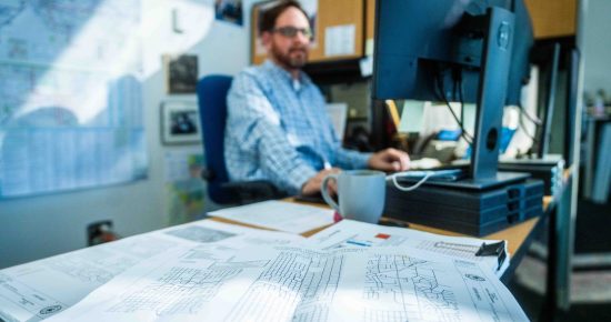 Plans on Desk with Man at computer behind.