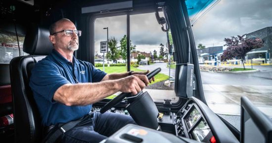 A bus driver wearing a blue shirt and glasses focuses on the road while driving. The bus interior is visible with a steering wheel, dashboard, and controls. Outside the window, a rainy day scene with a "one way" sign, trees, and buildings in the background.