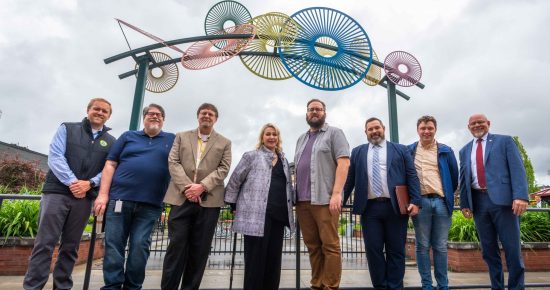 A group of eight people stands smiling under an outdoor sculpture made of circular and semi-circular materials in various colors. The sky is overcast, and they are dressed in business casual attire. The background features some greenery and a brick structure.