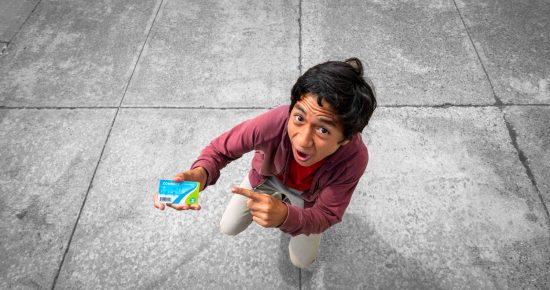 A young man kneels on the pavement, looking up excitedly, and points to a blue card he is holding in his other hand. The background features large concrete tiles.