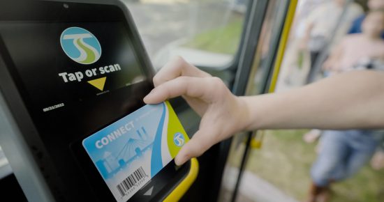 A person taps a blue Spokane Transit card on a black card reader labeled with a "tap or scan" sticker inside a bus, with passengers visible in the background.