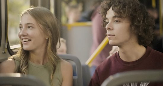 A young woman with long blonde hair wearing a green sleeveless top, and a young man with curly brown hair wearing a maroon sweatshirt sitting together on a bus. The woman is smiling and looking out the window while the man looks straight ahead.