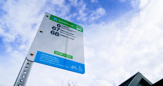 A Spokane Transit bus stop sign with route numbers 66 (EWU), 67 (Three Loop), and 68 (Cheney Loop) under a partly cloudy sky. The sign includes the stop number 3224 and contact information for real-time bus schedules.
