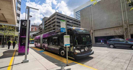 A modern city bus with "City Line" branding is at a bus stop on an urban street. The surrounding area features tall buildings, a bus shelter, and people walking. The sky is partly cloudy, and there is a signpost with bus route information near the bus.