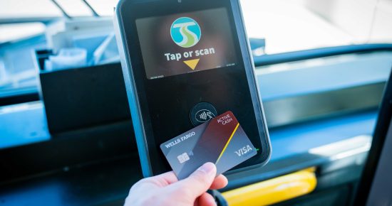 A hand holding a Wells Fargo Active Cash Visa credit card in front of a card reader on a bus. The card reader screen displays "Tap or scan" with an icon of a card being tapped.
