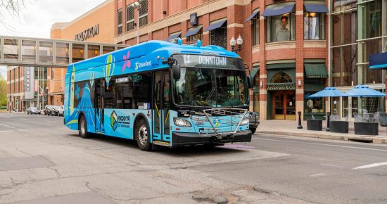 A city bus painted in bright blue with the text "Downtown" on the display is driving down a street lined with brick buildings. The buildings house various stores, including Gap and Anderson & Co. A pedestrian bridge connects two buildings above the street.