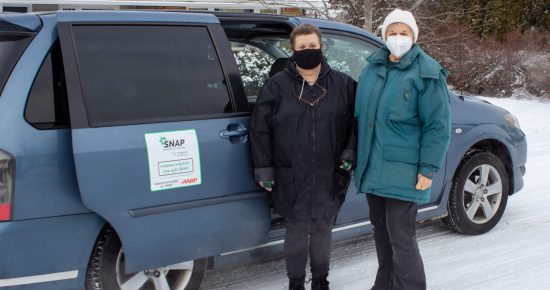 Two people, one wearing a black jacket and the other a blue jacket with masks, standing next to a Spokane Transit blue car in a snowy environment with a house in the background.