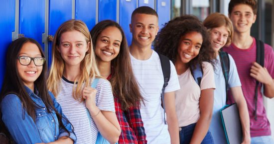 A group of seven diverse high school students stand in a hallway with their backs to a row of blue lockers. They smile at the camera, some holding notebooks and wearing backpacks. The mood is cheerful and friendly.