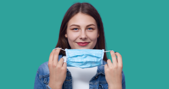 A young woman holding a face mask, smiling and looking at the camera. She wears a denim jacket, isolated on a turquoise background, promoting Spokane Transit's health safety initiatives.