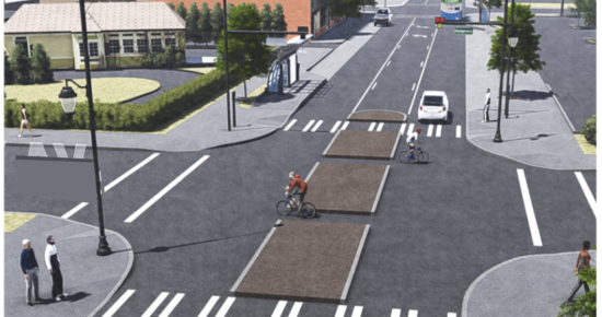 A city street with two bicyclists crossing at a crosswalk. Several cars travel on the street, and pedestrians wait at the corner. The area is lined with buildings, trees, and greenery, featuring bike lanes and designated crosswalks.