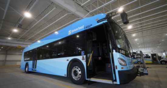 A light blue electric bus with the number E4002 is parked inside a spacious, well-lit transit facility. The bus door is open, revealing the interior. Branded with "Suburban Transit," it appears to be in a storage or maintenance area, adhering to the facility's style guide.