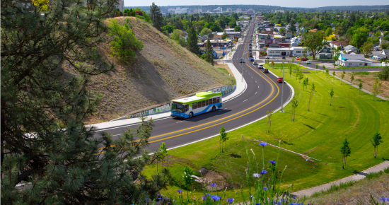 Aerial view of a city road with a Spokane Transit bus traveling, surrounded by lush greenery and a hill, with houses and trees in the background under a clear sky.