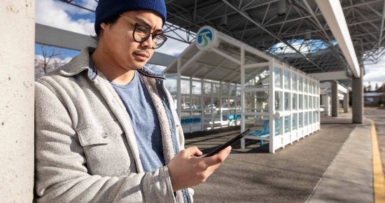 A person wearing a blue beanie, glasses, and a gray jacket leans against a concrete pillar while looking at a smartphone. They stand near a modern bus stop with clear panels and a blue sign overhead under a large metal roof structure.