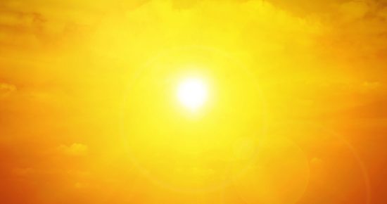 Bright yellow sun glowing intensely in a clear sky with a warm golden hue, diffusing light across the image. Few faint clouds are barely visible around the sun, enhancing the overall warmth and radiance. Sun flares create lens effects in the foreground.