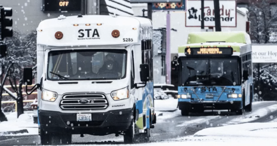 Two STA buses drive on a snowy street in a city. The bus in front is smaller and white, while the bus behind is larger and blue-green. Snow covers the ground and rooftops, and various business signs are visible in the background.